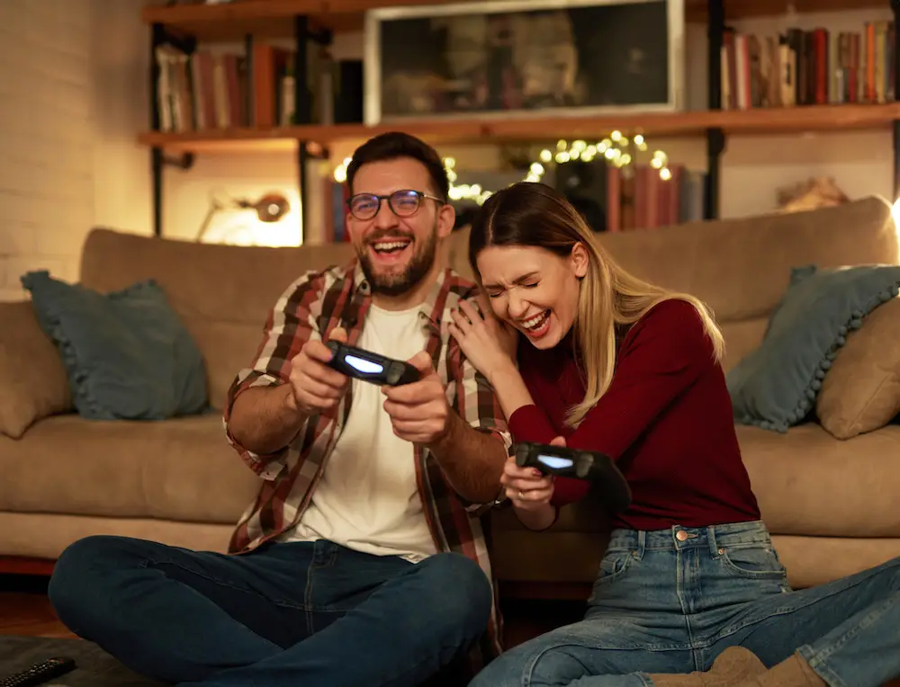 A young couple sitting in front of a couch laughing and playing PS4.